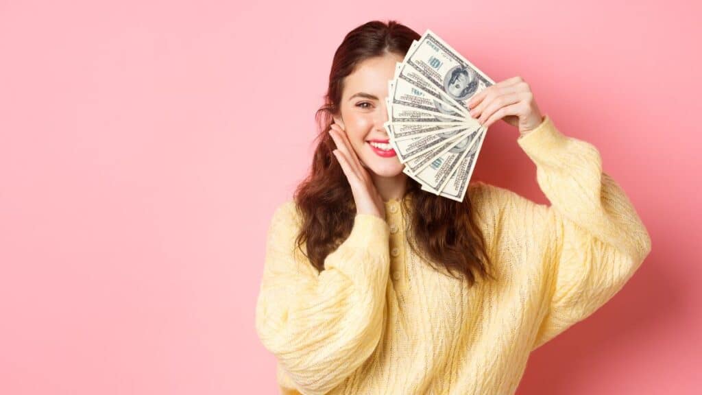 Woman holding a fan of cash in front of her face with a pink background