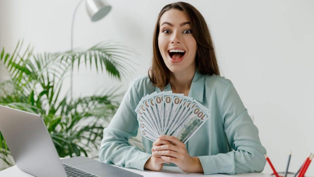 Woman holding a bunch of cash smiling in front of her laptop