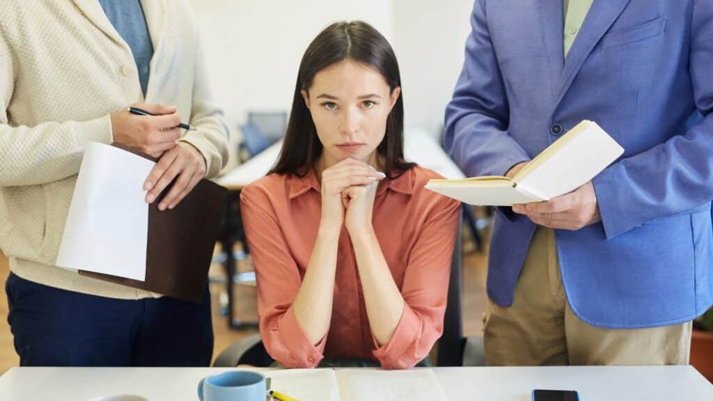 Woman sitting at a desk at work looking annoyed with coworkers standing next to her
