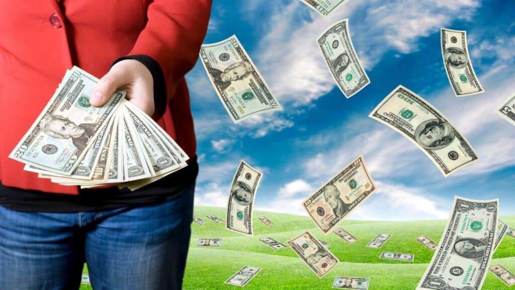 Woman holding cash in her hand in a field with cash flying around the sky
