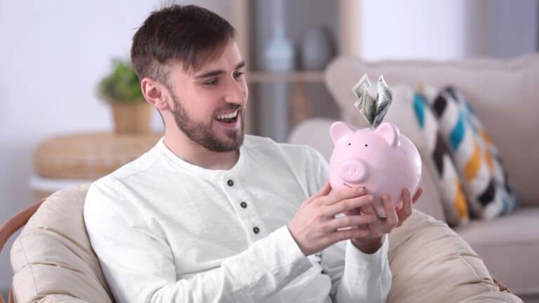 Man sitting on the couch smiling at a pink piggy bank with money