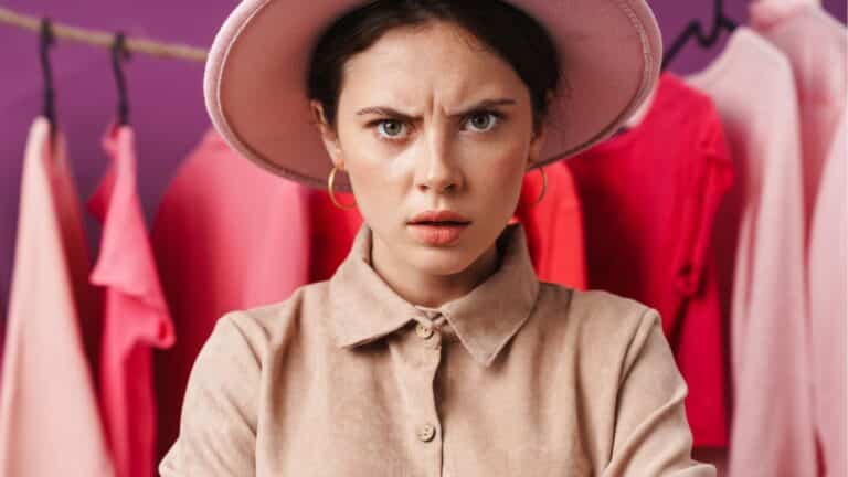 Woman in a store, wearing a hat looking annoyed while standing in front of a clothing rack