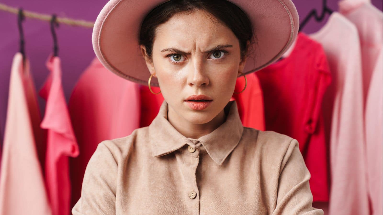 Woman in a store, wearing a hat looking annoyed while standing in front of a clothing rack