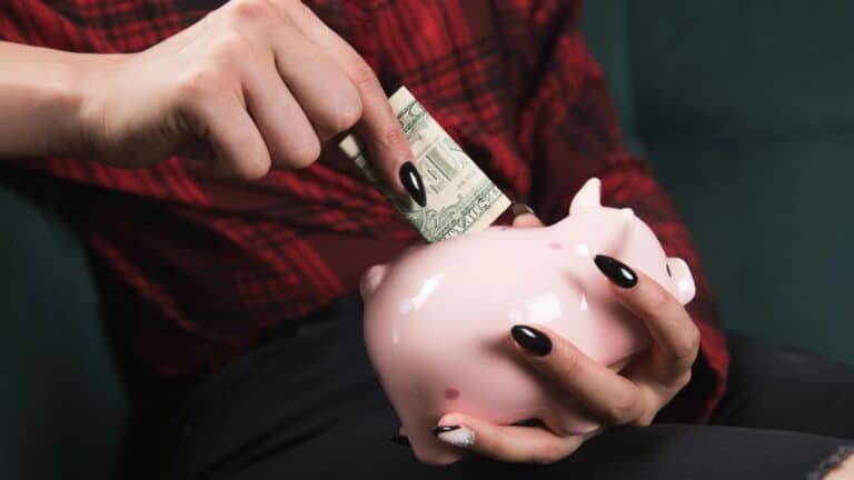 Woman with black fingernails putting money into a pink piggy bank