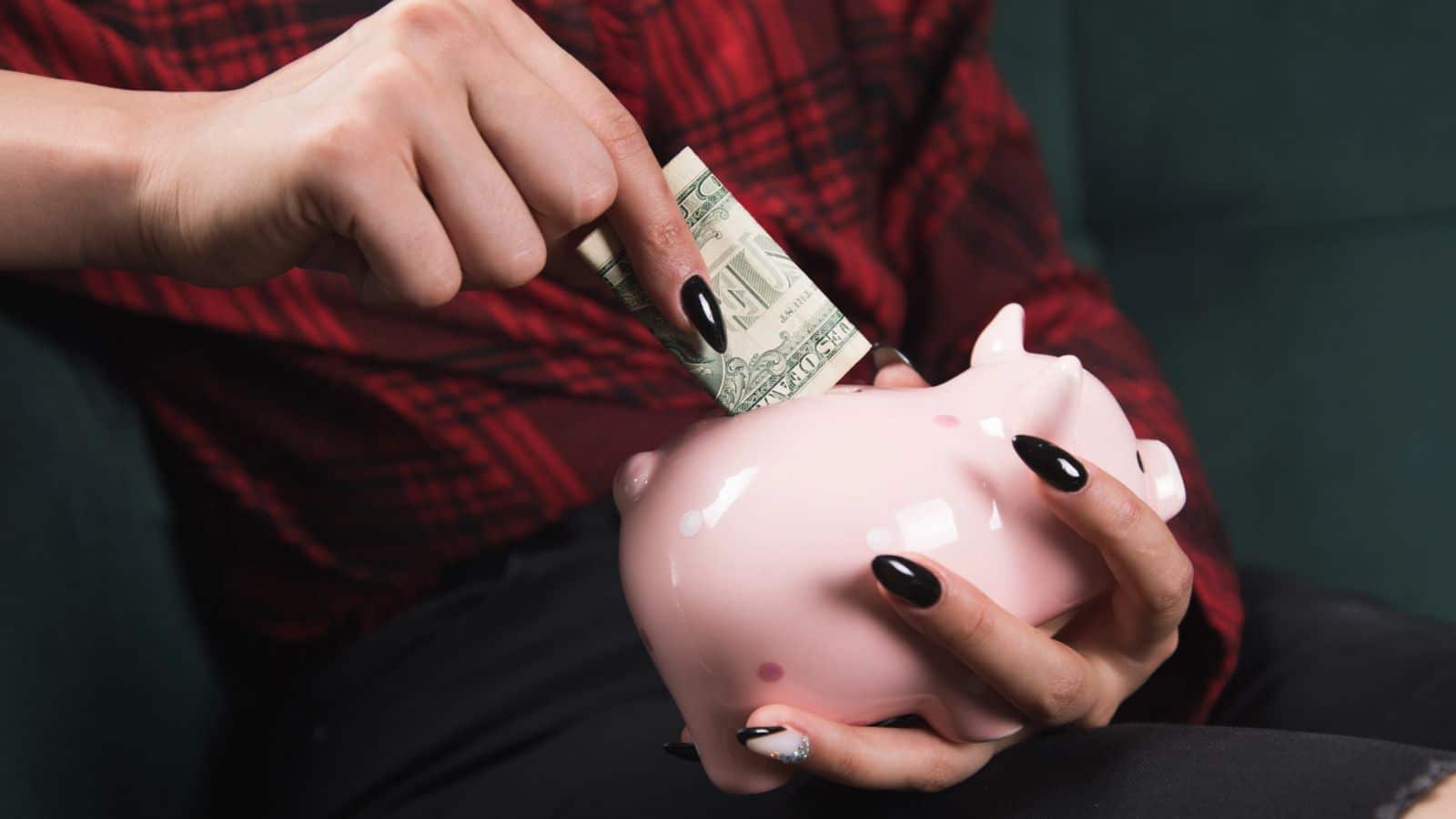 Woman with black fingernails putting money into a pink piggy bank