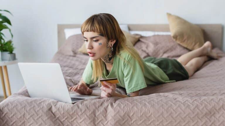 Gen Z woman laying on a bed shopping on her laptop with a credit card