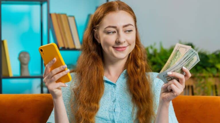 Redhead woman sitting on the couch holding her phone and a handful of cash