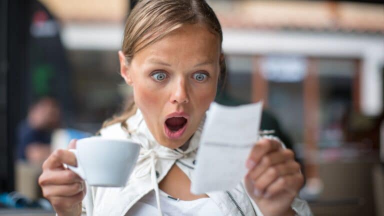 Woman holding a cup of coffee looking at a receipt with a shocked face