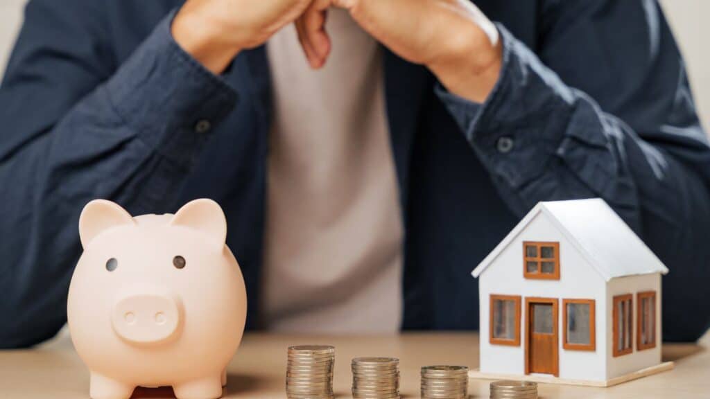 Man sitting with a piggy bank, a model of a house, and stacks of coins on a table
