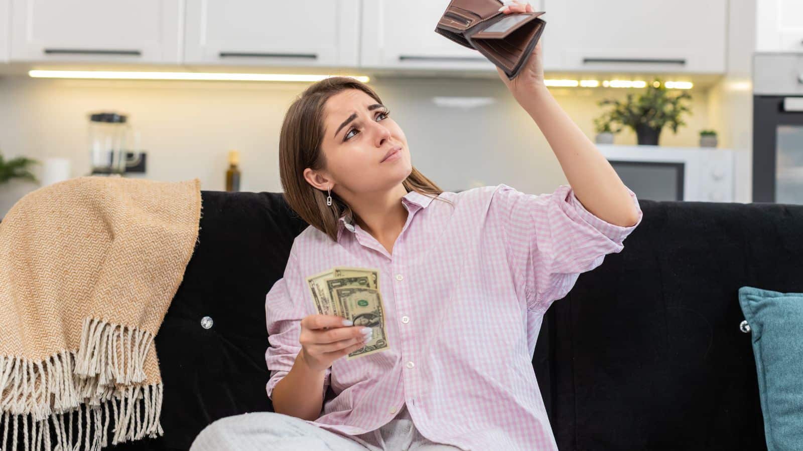 Woman sitting on couch holding money and looking at her empty wallet