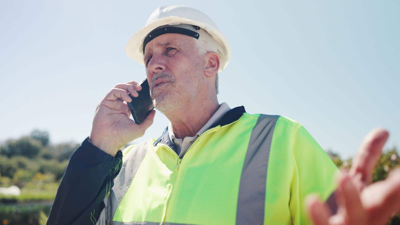 Man in bright vest and hard hat looking frustrated on the phone